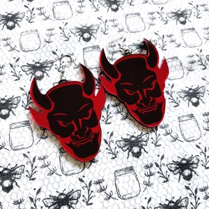 Könnte beinhalten: Ein Paar rot-schwarze Teufelsohrringe. Die Ohrringe zeigen ein detailliertes Teufelsgesicht-Design mit Hörnern und einem finsteren Grinsen. Die Ohrringe sind vor einem weißen Hintergrund mit einem schwarz-weißen Muster aus Bienen, Gläsern und Blättern angeordnet.