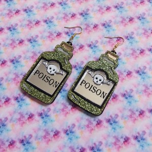 Peut inclure: Une paire de boucles d'oreilles pendantes en forme de flacons de potion avec une finition &agrave; paillettes vertes. Chaque flacon pr&eacute;sente une &eacute;tiquette argent&eacute;e avec le mot POISON imprim&eacute; en noir, un cr&acirc;ne blanc et des os crois&eacute;s, et des crochets dor&eacute;s.