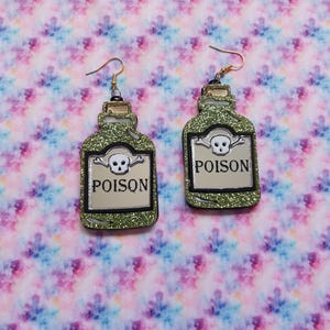Peut inclure: Une paire de boucles d'oreilles pendantes en forme de flacons de potion avec une finition &agrave; paillettes vertes. Chaque boucle pr&eacute;sente une &eacute;tiquette argent&eacute;e avec une ic&ocirc;ne de cr&acirc;ne et d'os crois&eacute;s au-dessus du mot POISON en lettres noires.