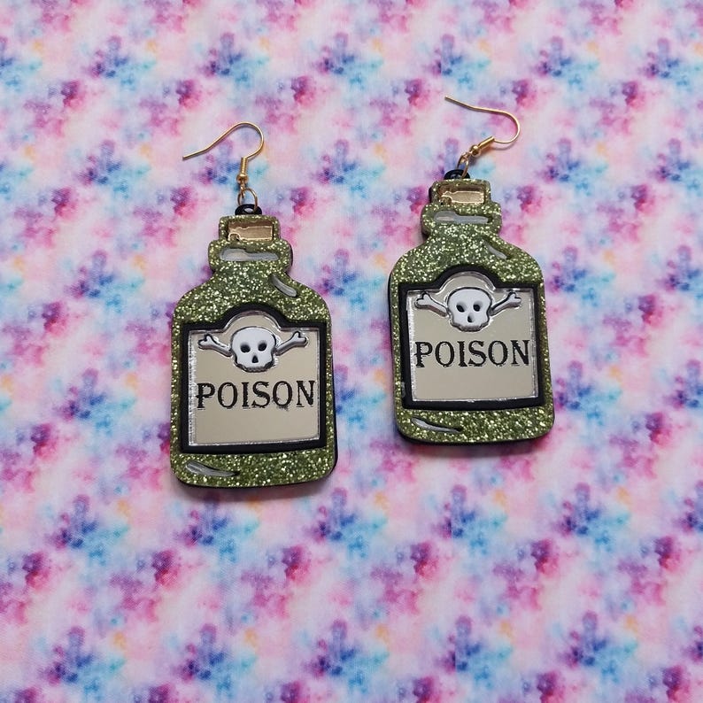 Peut inclure: Une paire de boucles d'oreilles pendantes en forme de flacons de potion avec une finition &agrave; paillettes vertes. Chaque boucle pr&eacute;sente une &eacute;tiquette argent&eacute;e avec une ic&ocirc;ne de cr&acirc;ne et d'os crois&eacute;s au-dessus du mot POISON en lettres noires.
