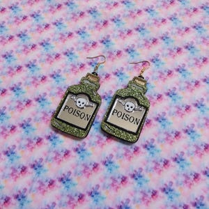 Peut inclure: Une paire de boucles d'oreilles pendantes en forme de flacons de potion. Les flacons sont verts avec une finition paillet&eacute;e et pr&eacute;sentent une ic&ocirc;ne de cr&acirc;ne et d'os crois&eacute;s blancs au-dessus du texte noir POISON. Chaque boucle a un crochet dor&eacute;.