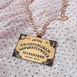 May include: Gold-colored Ouija board pendant necklace. The rectangular pendant features the alphabet, numbers, and the words "Yes," "No," and "Good Bye." The necklace has a thick gold chain.