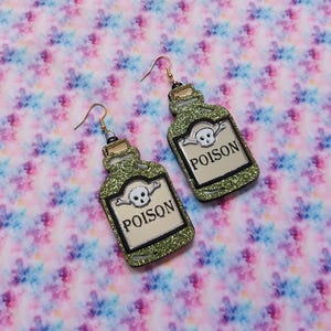 Peut inclure: Une paire de boucles d'oreilles pendantes en forme de flacons de poison vert &agrave; paillettes. Chaque flacon pr&eacute;sente une ic&ocirc;ne de cr&acirc;ne et d'os crois&eacute;s blanche au-dessus du texte noir POISON sur une &eacute;tiquette cr&egrave;me, avec des crochets dor&eacute;s.