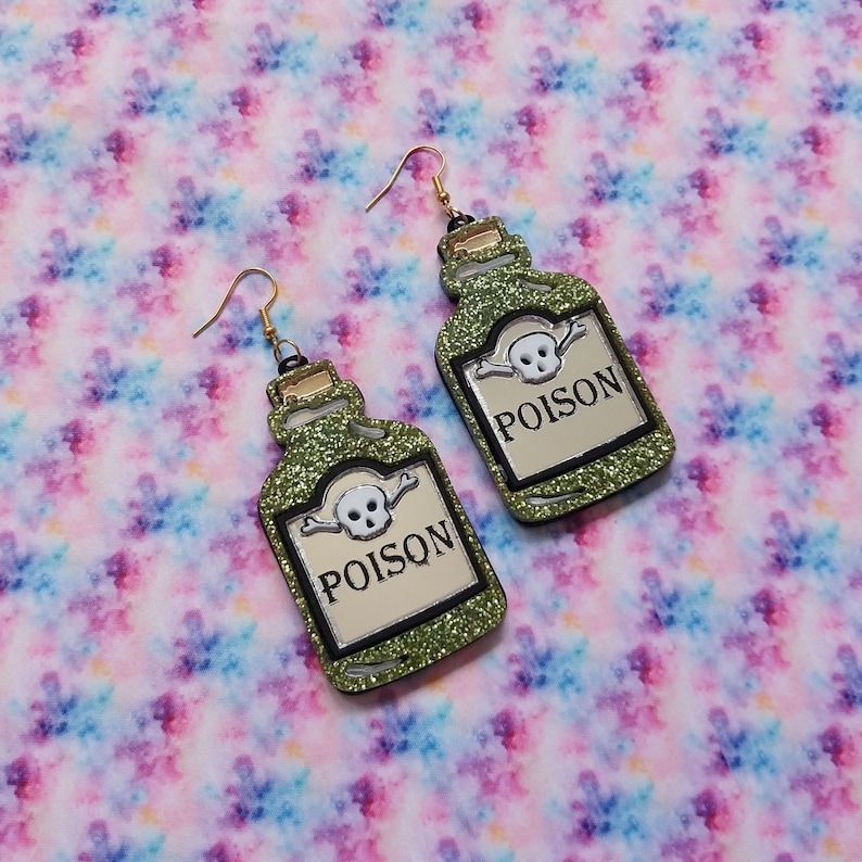 Peut inclure: Une paire de boucles d'oreilles pendantes en forme de flacons de poison vert &agrave; paillettes. Chaque flacon pr&eacute;sente une ic&ocirc;ne de cr&acirc;ne et d'os crois&eacute;s blanche au-dessus du texte noir POISON sur une &eacute;tiquette cr&egrave;me, avec des crochets dor&eacute;s.