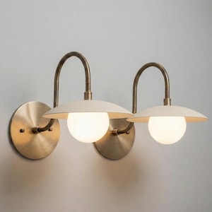 Könnte beinhalten: Zwei Wandleuchten mit Messing-Finish. Jede Leuchte hat einen cremefarbenen, tellerförmigen Schirm und eine kugelförmige, satinierte Glühbirne. Der gebogene Arm und die kreisförmige Rückplatte tragen zum Retro-Design bei.