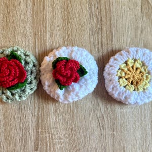Könnte beinhalten: Drei handgefertigte Häkeluntersetzer. Einer ist hellgrün mit einer roten Rose, einer ist weiß mit einer roten Rose und der dritte ist weiß mit einem gelb-orangenen Zentrum. Die Untersetzer sind rund und haben ein strukturiertes Aussehen.