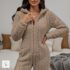 May include: A tan, hooded onesie with a full-length zip. The onesie features a textured, cable-knit design and long sleeves. The model is wearing gold necklaces and hoop earrings. The onesie is made of a soft, plush material.