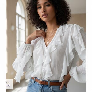 May include: White blouse with ruffled detailing and long sleeves. The blouse has a V-neck and button-up front. The model is wearing a brown belt and blue jeans. The blouse is made of a lightweight, textured fabric.