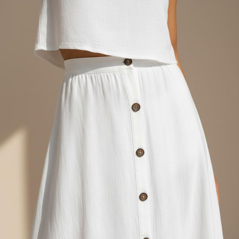 May include: A white skirt featuring a high waistband and a vertical row of five brown buttons down the center front. The skirt is paired with a matching white cropped top, both made of a textured fabric with a visible weave pattern.