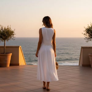 May include: A sleeveless white midi dress with a round neckline and a fitted bodice that transitions into a flowing, loose skirt. The garment is made of a lightweight, solid-colored fabric and is worn with simple flat sandals and a small woven bag.