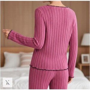 Może przedstawiać: Dwuczęściowy zestaw piżamy z prążkowanej dzianiny w kolorze magenta. Top z długim rękawem i spodnie mają pionową prążkowaną fakturę. Dekolt, mankiety i d&oacute;ł są wykończone czarną, falistą krawędzią. Modelka stoi tyłem do aparatu.