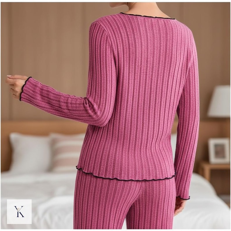 Może przedstawiać: Dwuczęściowy zestaw piżamy z prążkowanej dzianiny w kolorze magenta. Top z długim rękawem i spodnie mają pionową prążkowaną fakturę. Dekolt, mankiety i d&oacute;ł są wykończone czarną, falistą krawędzią. Modelka stoi tyłem do aparatu.