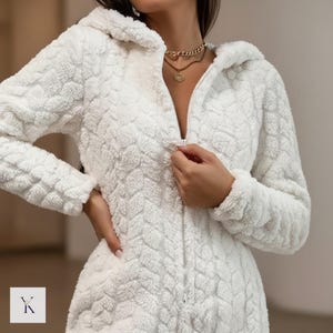 May include: A white, hooded, zip-up jumpsuit with a textured, chevron-like pattern. The plush fabric appears soft and warm. The jumpsuit has long sleeves and a full-length zipper. The image includes a small logo in the bottom left corner.
