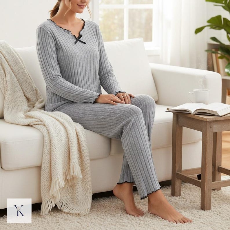 Może przedstawiać: Szary, prążkowany zestaw piżamowy z czarnym wykończeniem. Top z długim rękawem ma dekolt w serek z małą czarną kokardką. Spodnie mają pasującą prążkowaną fakturę. Modelka siedzi na białej sofie.