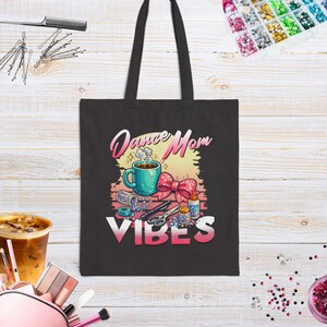 May include: Black tote bag with the words "Dance Mom Vibes" in a colorful design. The graphic includes a coffee cup, bow, and craft supplies. The bag has two black handles.