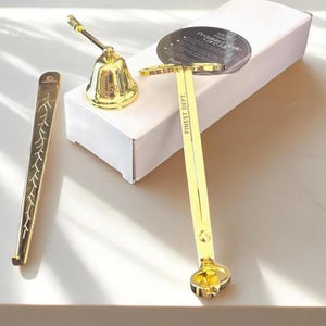 Puede incluir: Una colección de accesorios dorados para velas, que incluye una campana, un recortador de mecha y un apagador de velas. El recortador de mecha tiene el texto "FINEST DEPT." impreso. Los artículos están colocados sobre una caja blanca y una superficie clara.