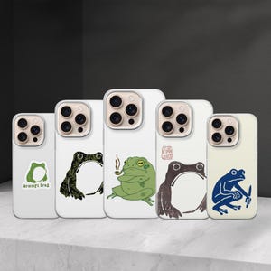 May include: A collection of white phone cases with unique frog illustrations. Designs include a green frog with a pipe, a blue frog with a knife, and a frog with the text "grumpt frog".