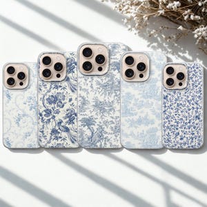 May include: Five phone cases with a blue and white toile pattern. The cases feature different designs, including floral, bird, and landscape motifs. The cases have a clear border around the camera.