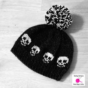 Könnte beinhalten: Schwarze Strickmütze mit weißem Bommel und Totenkopf-Designs. Die Mütze ist aus weichem, warmem Garn gefertigt und perfekt für kaltes Wetter.