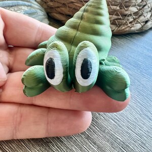 May include: A small, green, 3D-printed figurine shaped like a hermit crab with a spiral shell. The creature features two prominent, oval-shaped white eyes with large, vertical black pupils. The surface of the figurine displays visible horizontal layer lines.