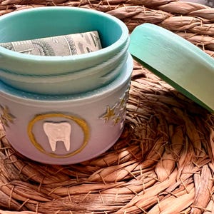 Op de afbeelding: Een kleine, ronde container in lichtpaars en blauw met een bijpassend deksel. De buitenkant heeft een wit tand-embleem in een gouden cirkel, omringd door kleine gouden sterren. De binnenkant is fel turquoise en bevat een gevouwen bankbiljet.