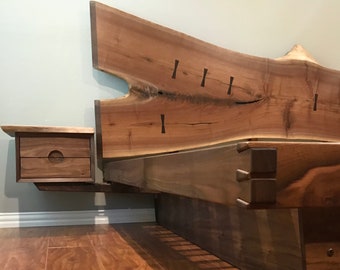 May include: A wooden bed frame with a live edge headboard and a matching nightstand. The bed frame is made of dark wood with a natural finish. The headboard is a single piece of wood with a live edge and a natural finish. The nightstand is a small, rectangular piece of wood with a drawer. The bed frame is sitting on a wooden floor.