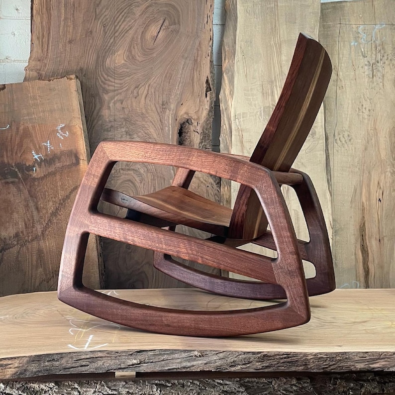May include: A dark brown wooden rocking chair with a unique, modern design. The chair is made of solid wood and has a comfortable, contoured seat. The chair is sitting on a large, wooden slab.