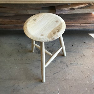 May include: A light wood stool with a round seat and three legs. The seat has a slightly curved edge and four small holes.
