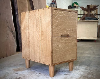 May include: A two-drawer wooden bedside table with a light wood finish. The drawers have a simple design with a small handle on each. The bedside table has four legs.