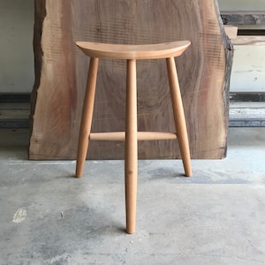 May include: A wooden stool with a curved seat and three legs. The stool is made of light wood and has a natural finish.