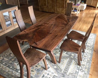 Könnte beinhalten: Ein handgefertigter Esstisch aus Holz mit einem Live-Edge-Design, zusammen mit vier passenden Stühlen. Tisch und Stühle sind aus dunkelbraunem Holz gefertigt, das die natürliche Maserung zeigt. Die Stühle haben geschwungene Rückenlehnen und Sitze.
