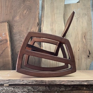 May include: A dark brown wooden rocking chair with a unique, modern design. The chair is made of solid wood and has a smooth, polished finish.