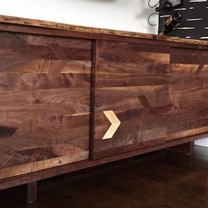 May include: A dark brown wooden cabinet with two sliding doors. The doors are made of wood with a geometric pattern and have a light brown arrow-shaped handle. The cabinet is sitting on a dark brown floor.