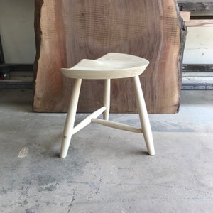 May include: A light wood stool with a curved seat and three legs. The stool is made of natural wood and has a simple, elegant design.
