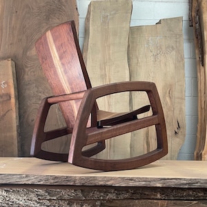 May include: A dark brown wooden rocking chair with a curved back and a wide, open seat. The chair is made of solid wood and has a natural finish. The chair is sitting on a wooden surface.