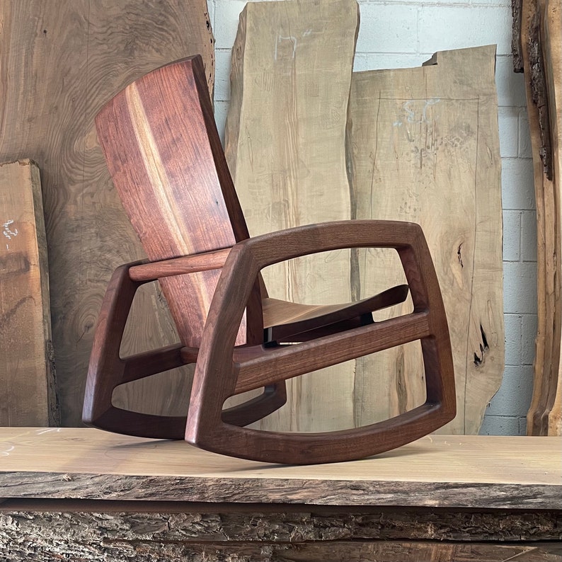 May include: A dark brown wooden rocking chair with a curved back and a wide, open seat. The chair is made of solid wood and has a natural finish. The chair is sitting on a wooden surface.