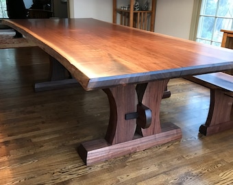 May include: A large, live-edge walnut dining table with a natural wood finish. The table has two sturdy, curved legs made of walnut wood. The table is paired with a matching bench.