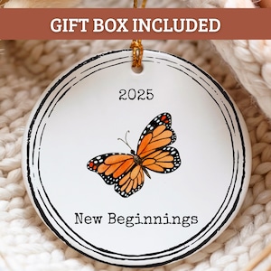May include: White ceramic ornament with a black border, featuring a monarch butterfly with orange and black wings. The year "2025" and the words "New Beginnings" are printed on the ornament. A gold string is attached for hanging.