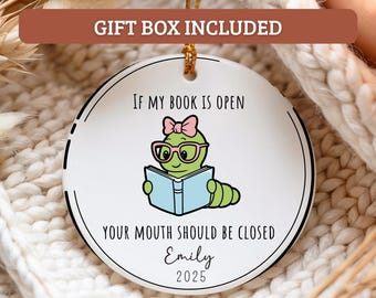 May include: A white ceramic ornament with a black border and gold string. The ornament features a cartoon worm wearing glasses and a bow, reading a book. Text reads "IF MY BOOK IS OPEN YOUR MOUTH SHOULD BE CLOSED Emily 2025".