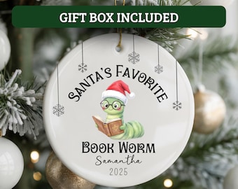 May include: A white ceramic Christmas ornament with a cartoon bookworm wearing a Santa hat and glasses, reading a book. The ornament reads "Santa's Favorite Book Worm" with a name and year. Includes a gold string for hanging.