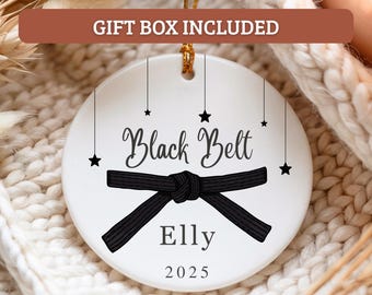 May include: A white ceramic ornament with a black belt design, the words "Black Belt" and the name "Elly" are written in black script. The year "2025" is printed below the name. Black stars dangle from the top.