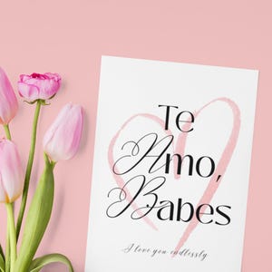 May include: A white card with the words "Te Amo, Babes" in black script, with a pink heart outline. The phrase "I love you endlessly" is written below. Pink tulips are arranged on the left side of the image.