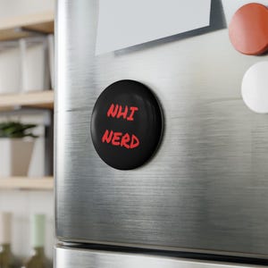 Puede incluir: Un imán redondo negro con las palabras "NHI NERD" en rojo, exhibido en la puerta de un refrigerador de acero inoxidable. El diseño del imán es simple y audaz.