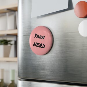 Puede incluir: Un imán redondo rosa con las palabras "Yarn Nerd" en negro, con letras manuscritas. El imán está adherido a la puerta de un refrigerador de acero inoxidable. Se ven otros imanes.