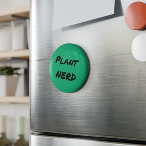 Puede incluir: Un imán redondo verde con las palabras "PLANT NERD" en negro, con letras de estilo manuscrito. El imán está adherido a la puerta de un refrigerador de acero inoxidable. Otros imanes son visibles.
