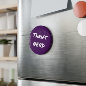 Puede incluir: Un imán redondo morado con las palabras "Thrift Nerd" en blanco, con letras de estilo manuscrito. El imán está adherido a la puerta de un refrigerador de acero inoxidable. Se ven otros imanes.