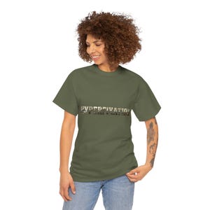 Könnte beinhalten: Olivgrünes T-Shirt mit dem Wort "HYPERFIXATION" in einem Camouflage-Stil-Schriftzug über der Brust. Das Shirt hat kurze Ärmel und einen Rundhalsausschnitt. Das Model trägt blaue Jeans.