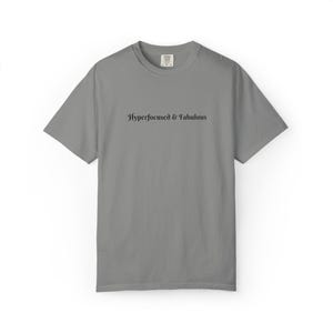 Op de afbeelding: Een grijs t-shirt met korte mouwen en een ronde hals, voorzien van de tekst Hyperfocused & Fabulous in een zwart schreeflettertype in het midden van de borst. Het kledingstuk heeft een standaard pasvorm met een geribbelde halslijn.
