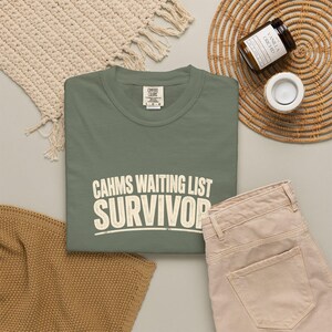May include: A sage green crewneck t-shirt featuring the cream-colored text "CAHMS WAITING LIST SURVIVOR" printed in a distressed, bold, sans-serif font across the chest. The shirt has a Comfort Colors brand label at the inner neckline.