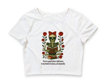 Può includere: Un top corto bianco a maniche corte con la grafica di uno scheletro che indossa un fiocco rosso e occhiali da sole, seduto a gambe incrociate mentre legge un libro circondato da rose rosse. Il testo recita: I'm in a good place right now, in my book of course, not mentally.